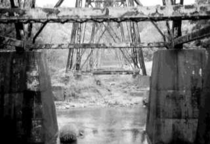 Black and white photo of an old iron railway bridge over a river, evoking unsettling Wikipedia article scenes.