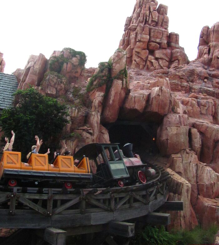 Roller coaster train with passengers raising hands, approaching rocky mountain tunnel, evoking scary Wikipedia article vibes.