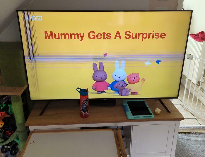 TV screen with damaged display showing a kids' show titled Mummy Gets A Surprise, in a family living room with toys visible nearby.