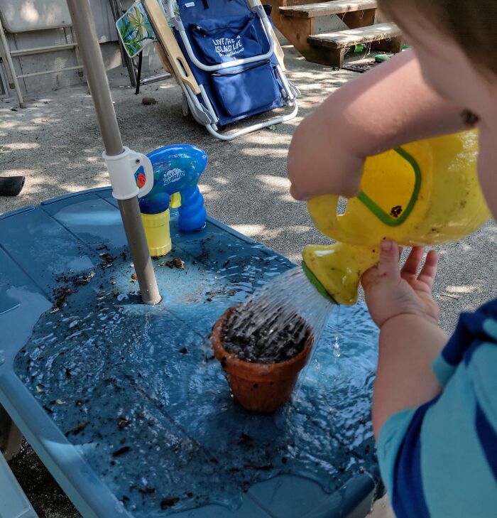 Niño regando una maceta con regadera amarilla, ejemplificando un día intenso para padres y madres.