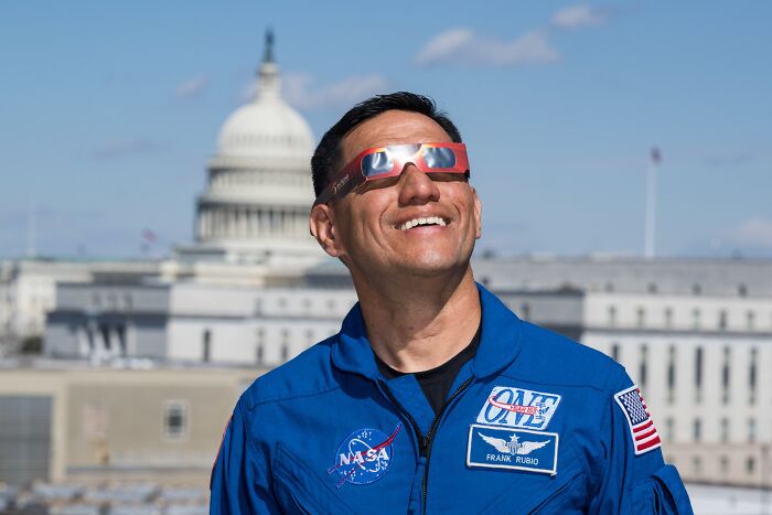 Astronaut Frank Rubio wearing NASA flight suit and eclipse glasses with Capitol building in background under blue sky.
