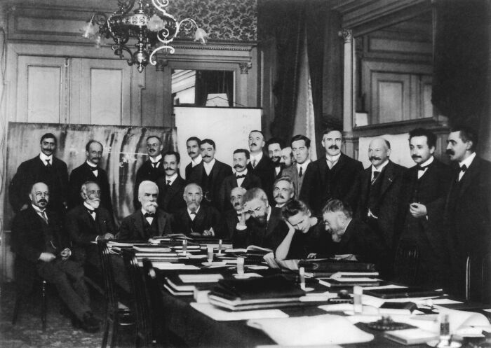 Black and white historical photo of a group of men in suits around a table, related to Marie Curie facts and discoveries.