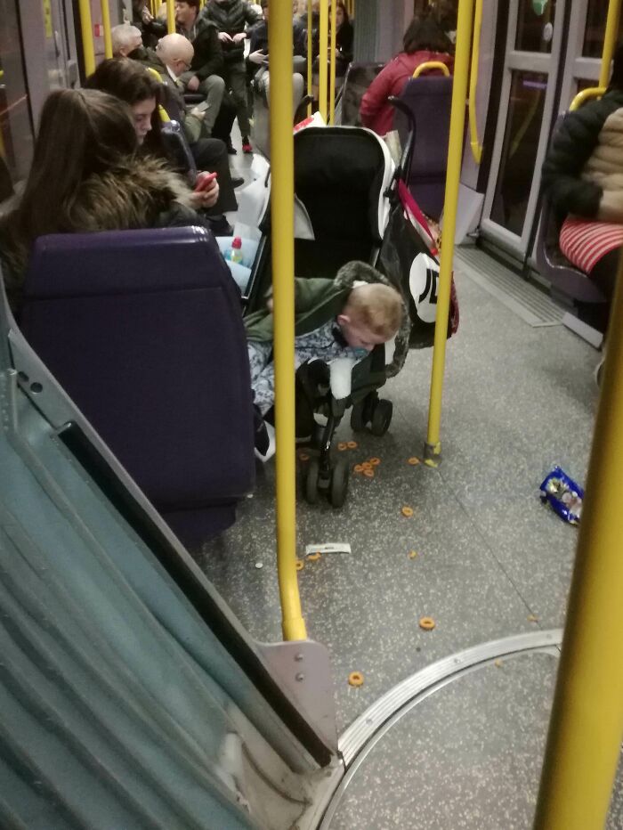 Child spilling snacks from stroller on crowded public transport with passengers seated and standing nearby.