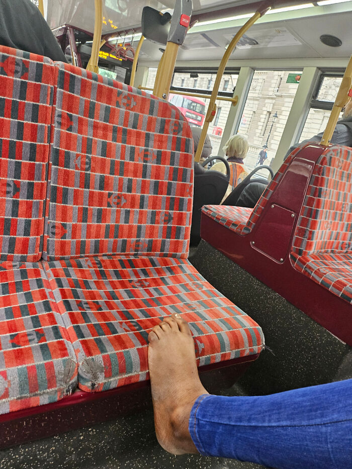A public transport passenger resting a bare foot on a red patterned bus seat, showing rude behavior.