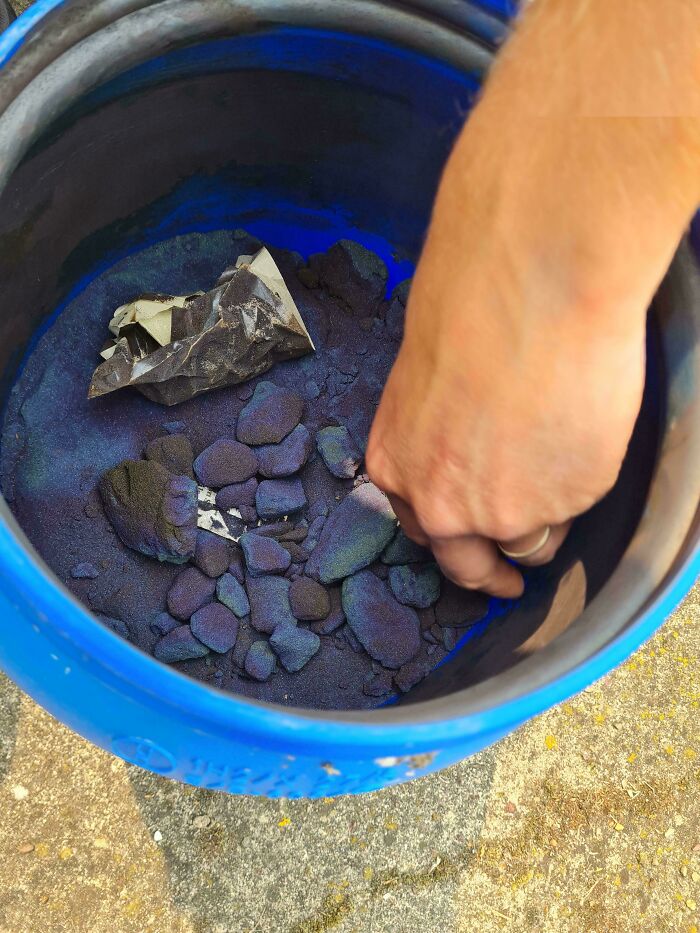 Hand touching mysterious dark purple stones and powder inside a blue container in a mystery objects recognition group.