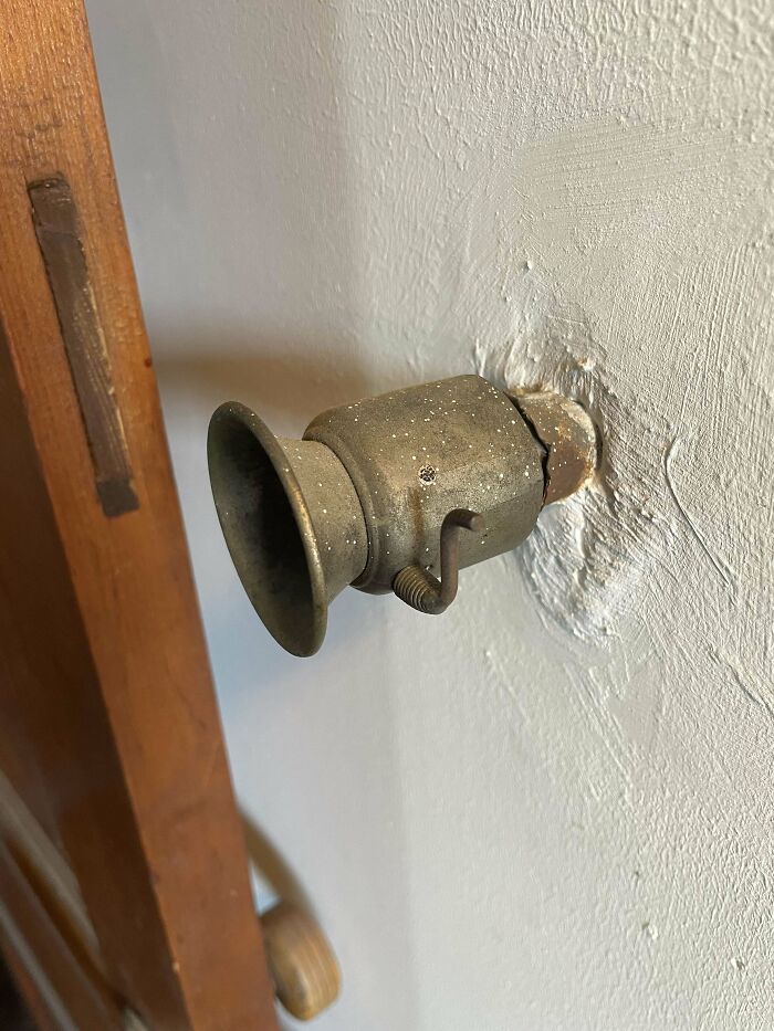 Old metal mystery object mounted on wall with a tapered, horn-like opening and a small lever on the side.