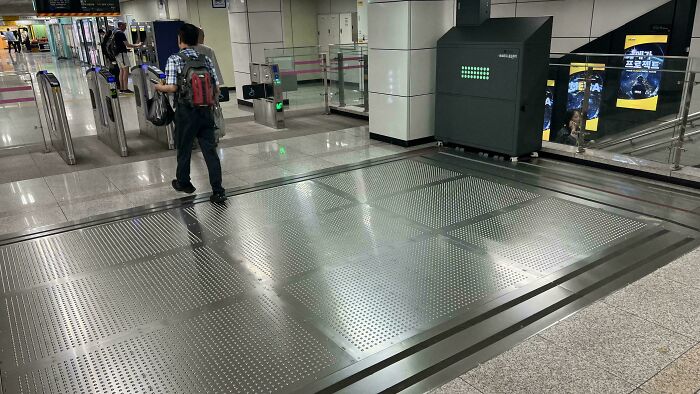 Man with backpack standing near a large perforated metal floor panel in a subway station mystery objects recognition context.