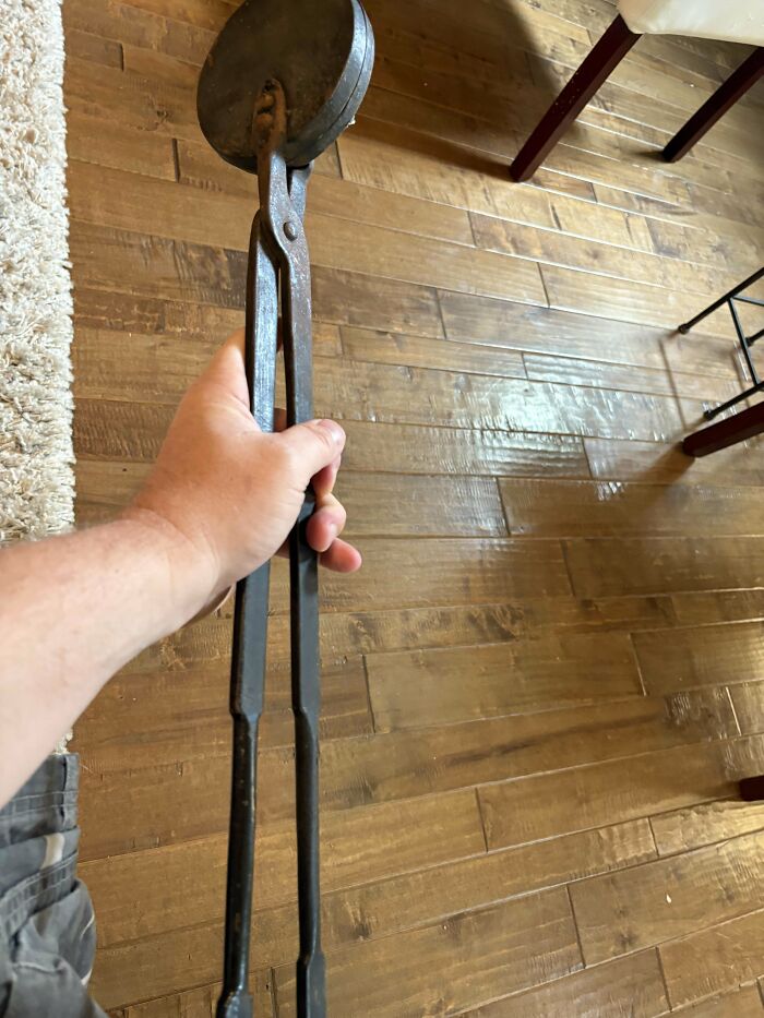 Hand holding a rusty mystery object with long handles and a round flat end on wooden floor indoors.