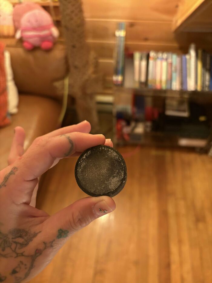 Hand holding a small, round, black mystery object indoors with wooden floor and shelves in the background.