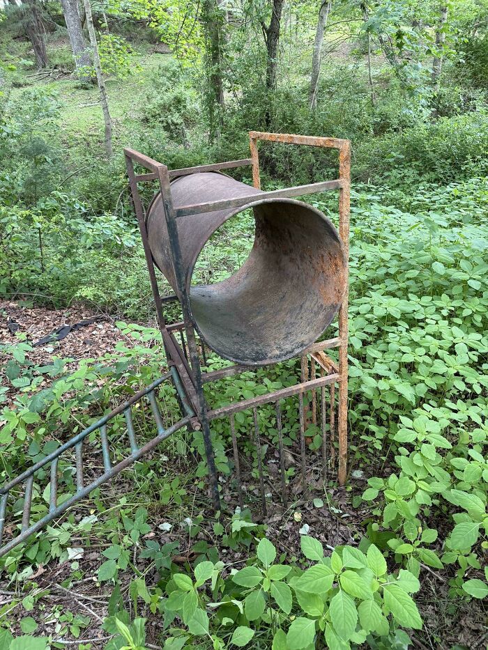 Rusty metal mystery object with a large hollow cylinder and ladder frame in overgrown green forest setting.