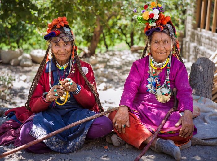 Two elderly women from around the world wearing traditional colorful clothing and floral headpieces smiling outdoors.