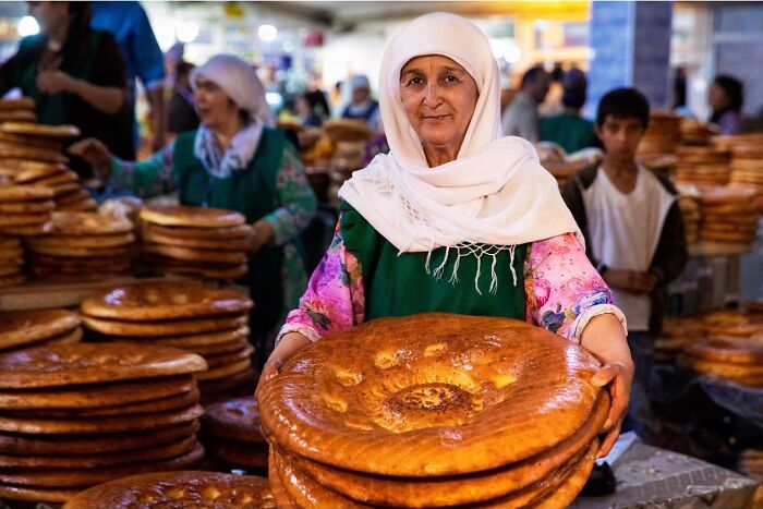 Elderly woman wearing a hijab holding traditional large bread at a busy market, showcasing beauty beyond age.