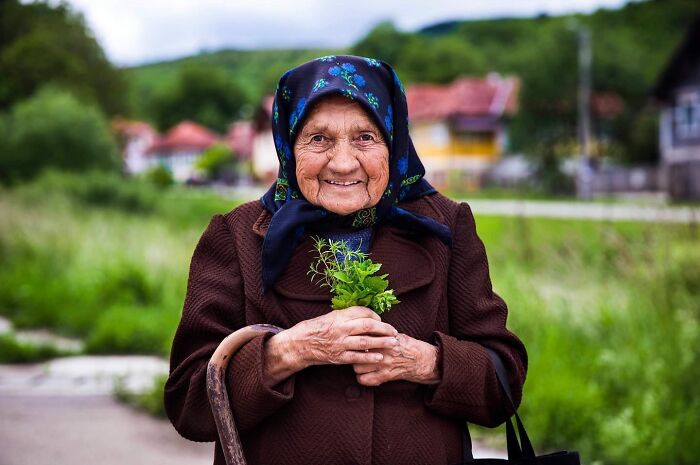Elderly woman wearing a headscarf smiling outdoors, holding fresh herbs, showcasing natural beauty and grace.