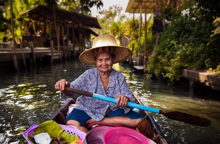 Elderly woman paddling boat on canal, wearing traditional hat and smiling, showcasing beauty and culture worldwide.