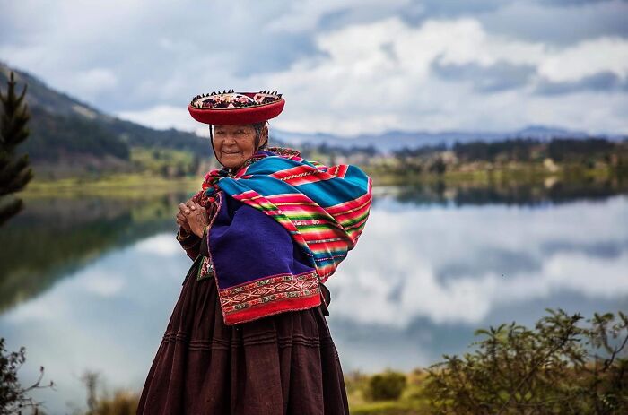 Elderly woman in traditional colorful clothing posing by a lake, showcasing timeless beauty from around the world.