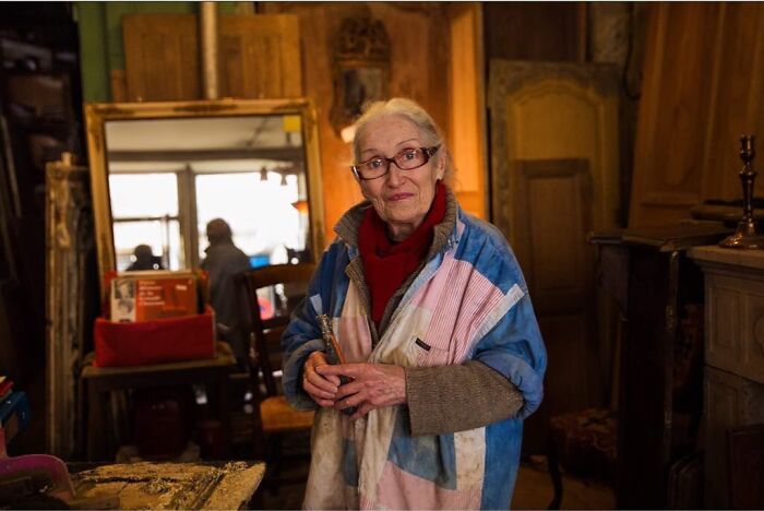Elderly woman with glasses in colorful smock holding paintbrush in cozy artist studio showcasing ageless beauty.
