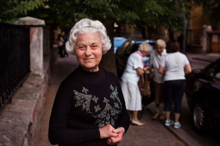 Elderly woman with white hair smiling outdoors, wearing a black sweater with butterfly designs, showcasing timeless beauty.