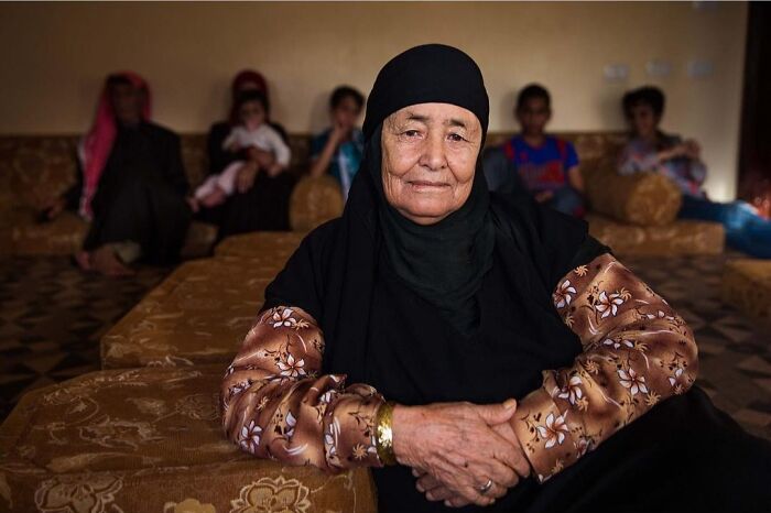 Elderly woman wearing a black headscarf sitting indoors with family in the background, showcasing beauty without expiration date.