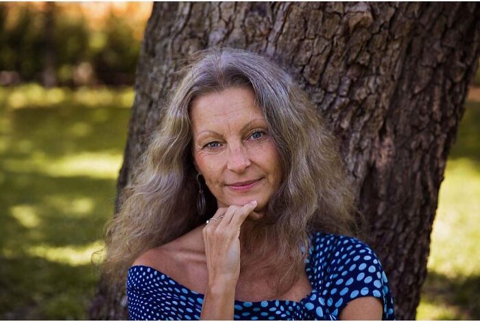 Elderly woman with long wavy hair in a blue polka dot dress sitting outdoors, embodying timeless beauty and grace.