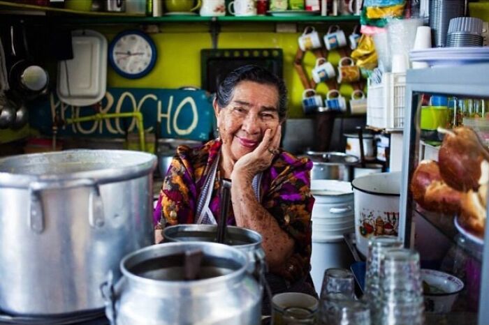 Elderly woman in a colorful shirt smiling warmly while sitting in a cozy kitchen surrounded by cooking pots and utensils.