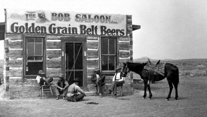 Vintage cowboy photograph showing men relaxing outside a rustic saloon in the wild west with a saddled horse nearby.