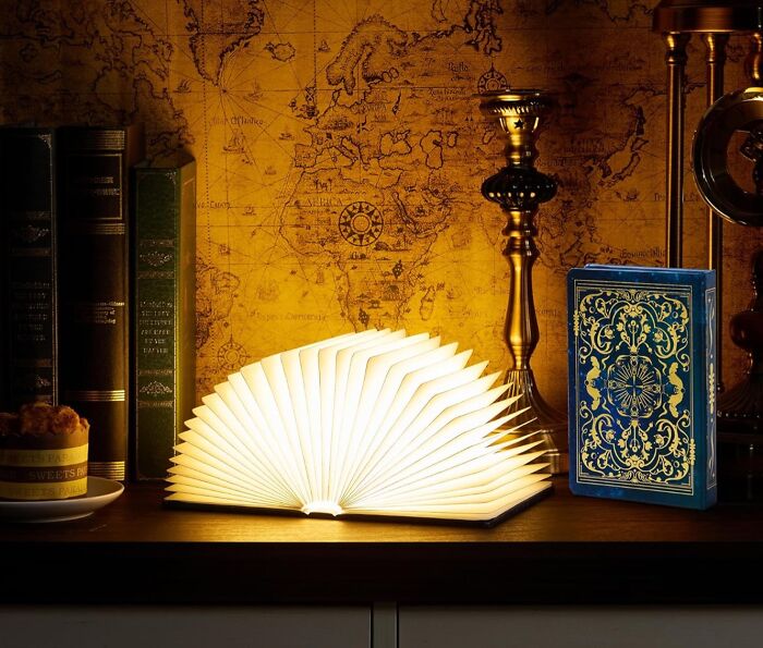 Glowing book lamp shaped like an open fan on a wooden shelf with antique map background and decorative arty finds.