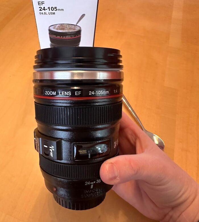 Hand holding a creative spirit inspired camera lens coffee mug with a spoon and packaging on wooden table.