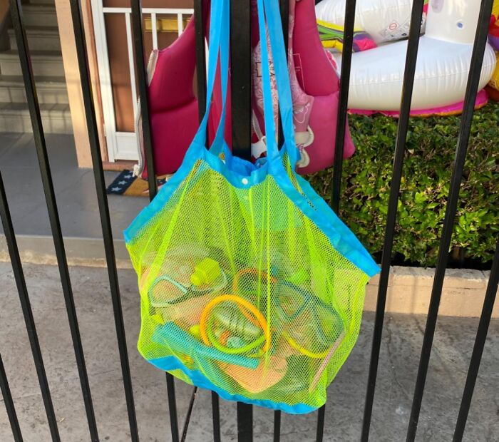 Bright blue and yellow mesh beach bag hanging on black fence, filled with sun and sand essentials for a perfect beach day.
