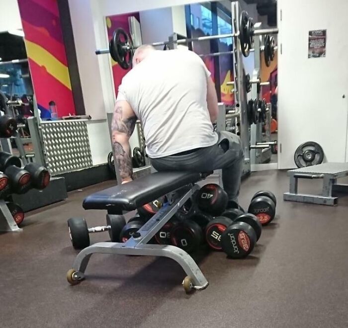 Man sitting on gym bench surrounded by dumbbells, illustrating annoying gym moments disrupting workouts.