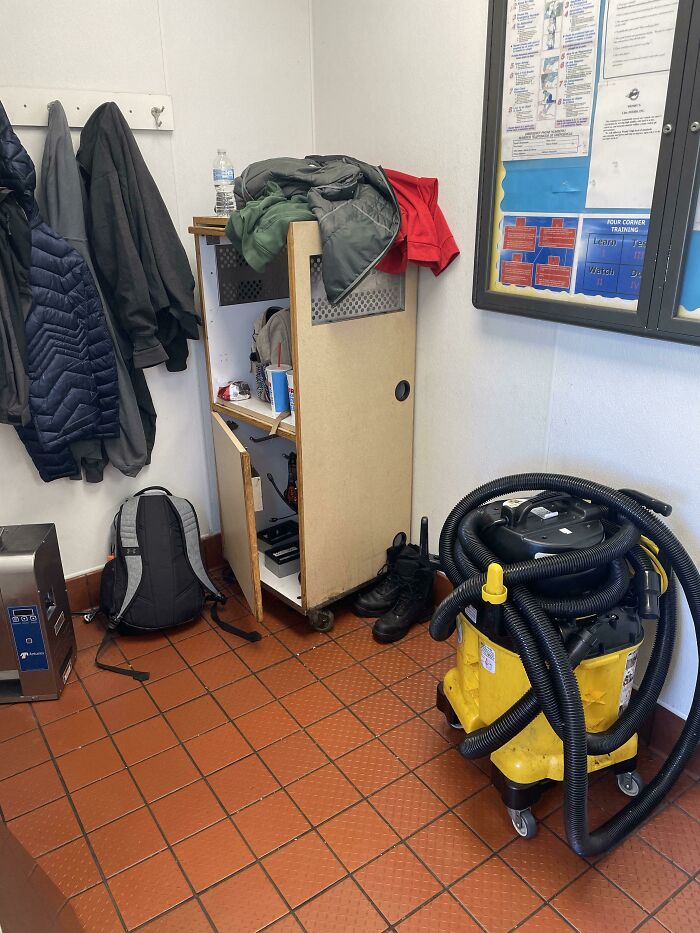 Cluttered employee breakroom with coats, backpack, broken cabinet, and tangled vacuum in toxic work environment.