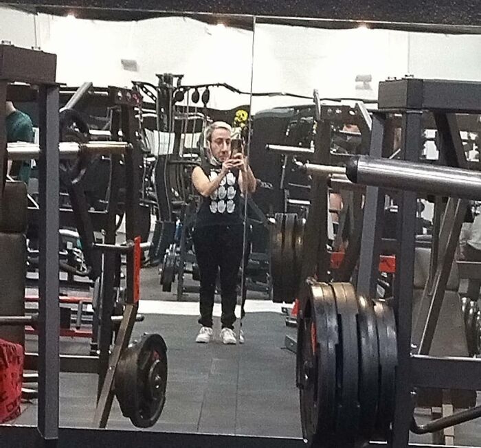 Woman taking a mirror selfie in a crowded gym filled with weightlifting equipment and heavy plates around.