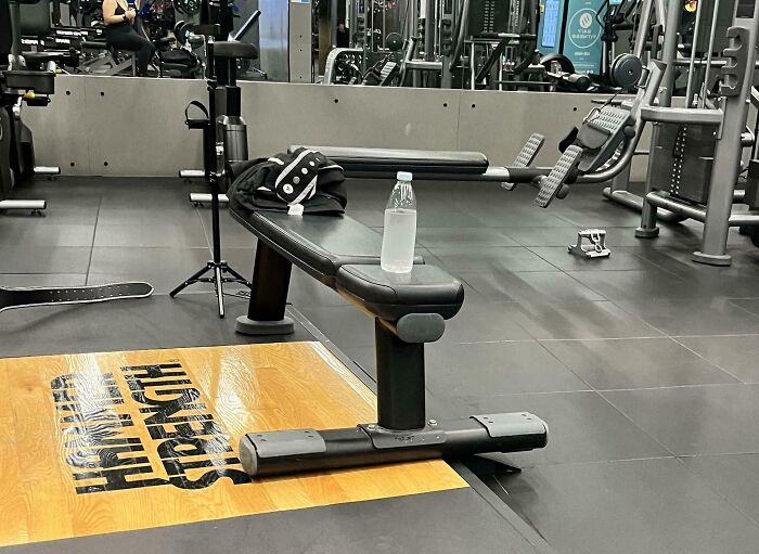Empty gym bench with a water bottle and towel, surrounded by workout machines and weights in a fitness center.