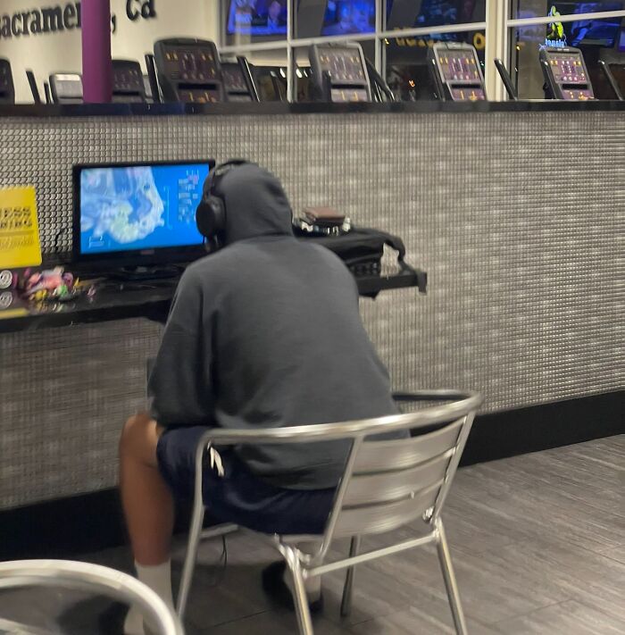 Person in a gym wearing headphones and a hoodie sitting on a metal chair playing on a screen during an annoying gym moment.