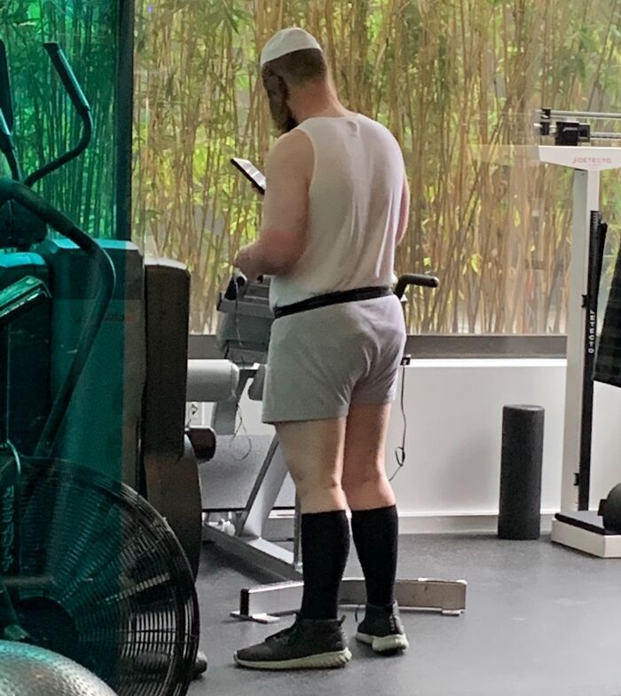 Man in gym shorts and tank top using phone while standing near exercise equipment in an indoor gym setting with plants outside.