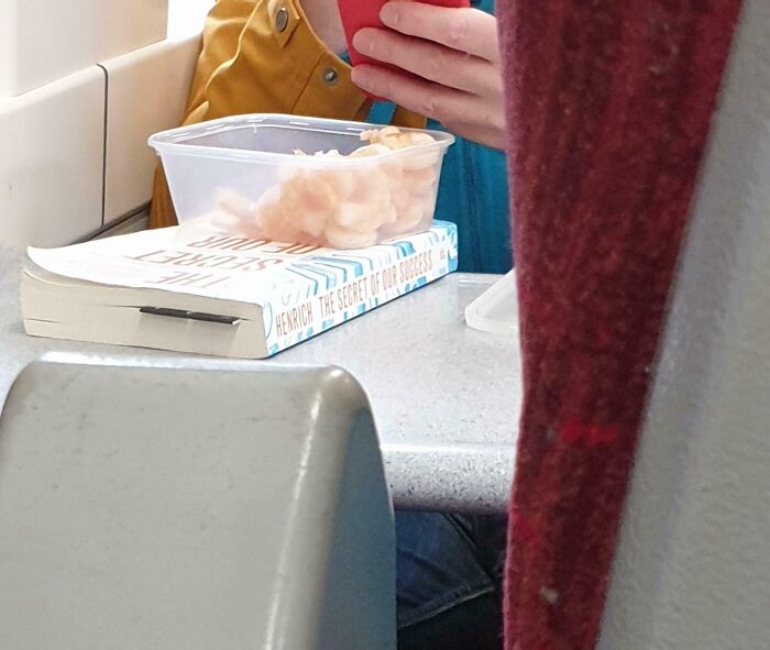 Passenger eating shrimp from a container on public transport, with a book placed on the table nearby.