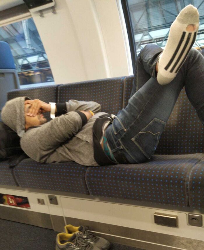 Passenger lying across multiple seats with feet up, wearing socks with holes, on public transport during commute.