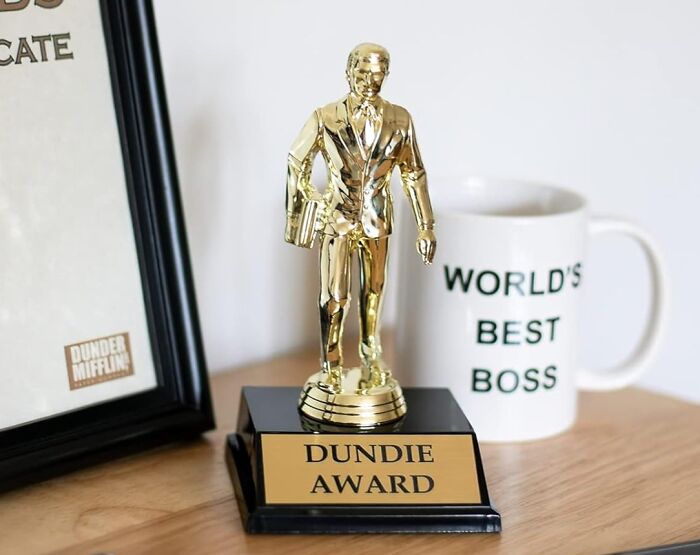 Desk with a Dundie Award trophy, framed certificate, and a white mug reading World's Best Boss, showcasing quirky office supplies.
