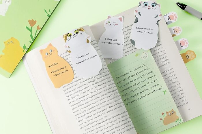 Cat-shaped office supplies including sticky notes and page markers arranged on an open book and green background.