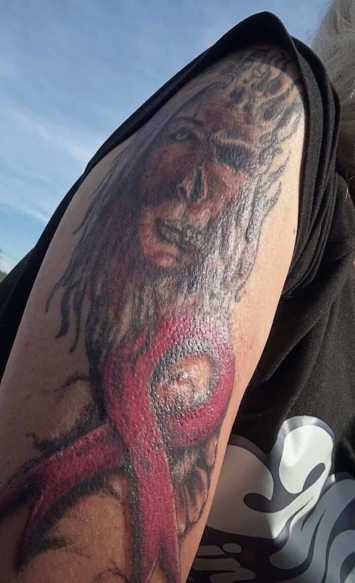 Bad tattoo on upper arm showing a distorted face and an unclear red shape, illustrating hilariously bad tattoos and inkshaming.