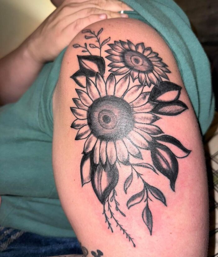 Black and gray sunflower tattoo on upper arm showcasing detailed leaves and petals in a hilariously bad tattoo style.