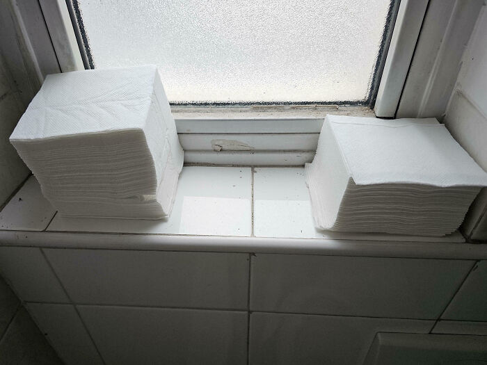 Two uneven stacks of white paper towels on a grimy window ledge in a toxic work environment restroom.