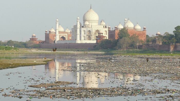 A famous landmark, the Taj Mahal, reflected in shallow water from an unexpected angle showing the surrounding landscape.