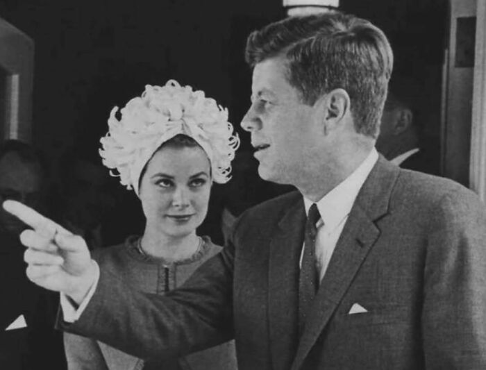 Black and white photo of a man in a suit pointing while a woman with a decorative hat looks on, history theme.