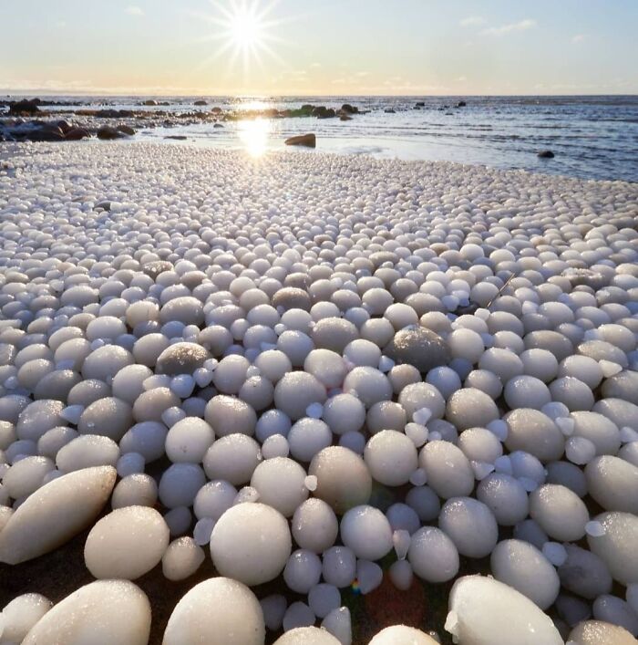 Unreal photograph of countless ice balls on a beach under the bright sun by the ocean shore at sunrise.