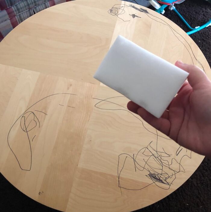 Hand holding a multi-tasking magic eraser cleaning marker scribbles off a wooden home table surface.