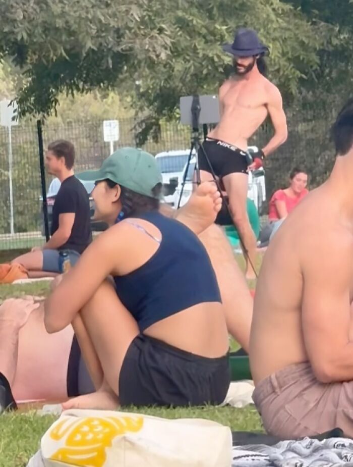 Shirtless influencer wearing a hat and black shorts performing a stunt in a park, attracting attention from seated people.