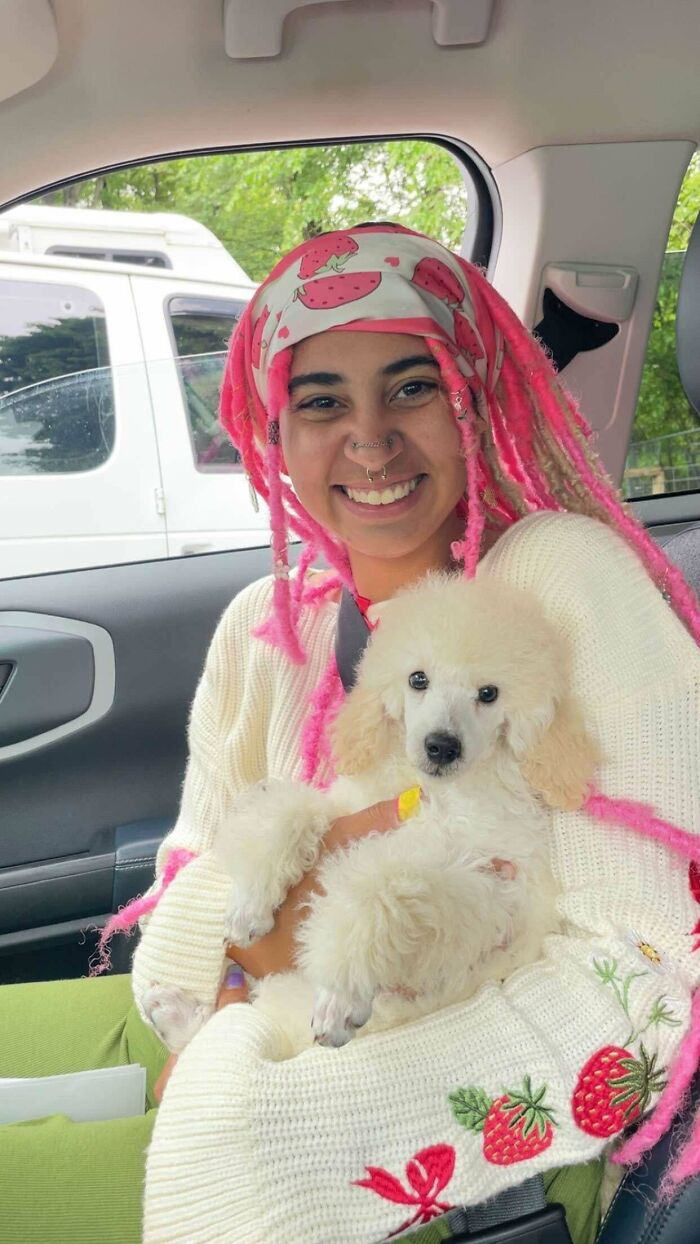 Smiling person with pink hair holding a fluffy white puppy in a car, capturing a wholesome adoption moment.