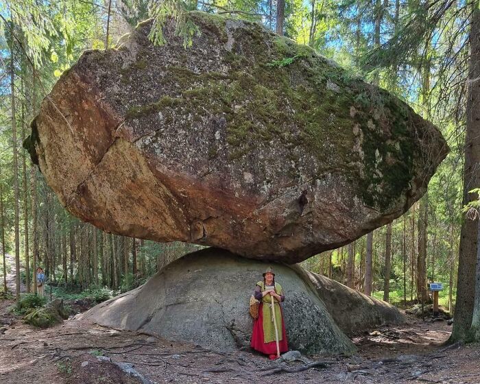 Woman standing under a massive balanced rock formation in a forest, showcasing one of the most unreal photographs.