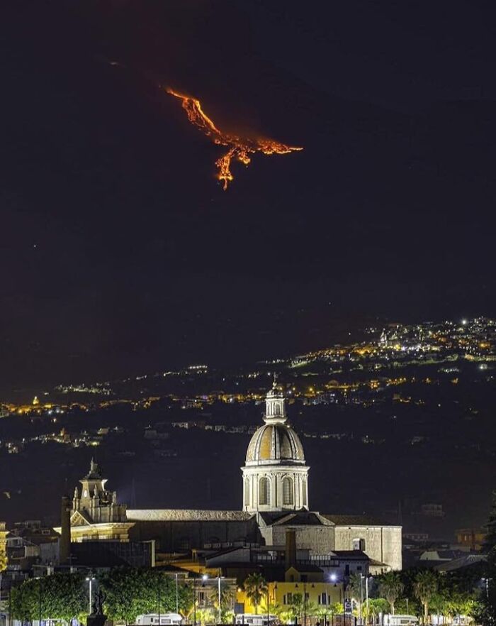 Night view of an unreal photograph showing a glowing lava flow over a dark mountain above a lit cityscape with a domed building.