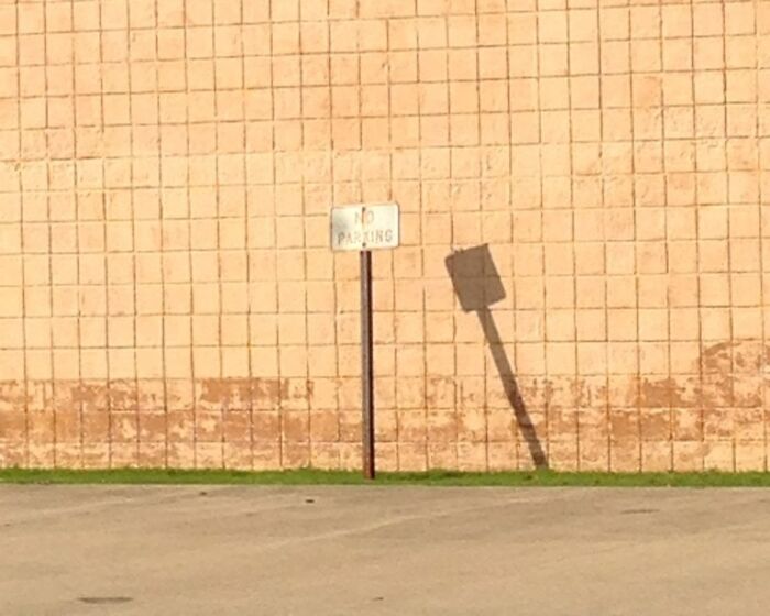 Shadow of a no parking sign casting a tilted, distorted shape on a textured brick wall in an empty parking lot.